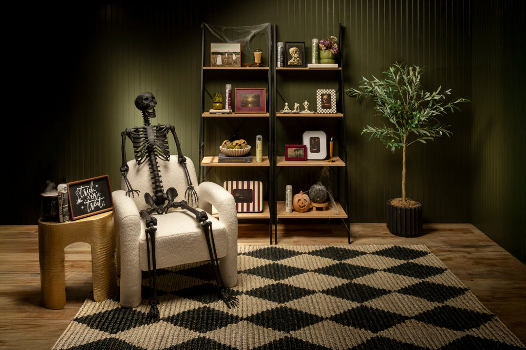 Dimly lit room with black skeleton sitting on white chair, checkerboard rug underneath. Table with "trick or treat" sign. Book shelves with assorted frames and Halloween decor. Faux potted tree in corner.
